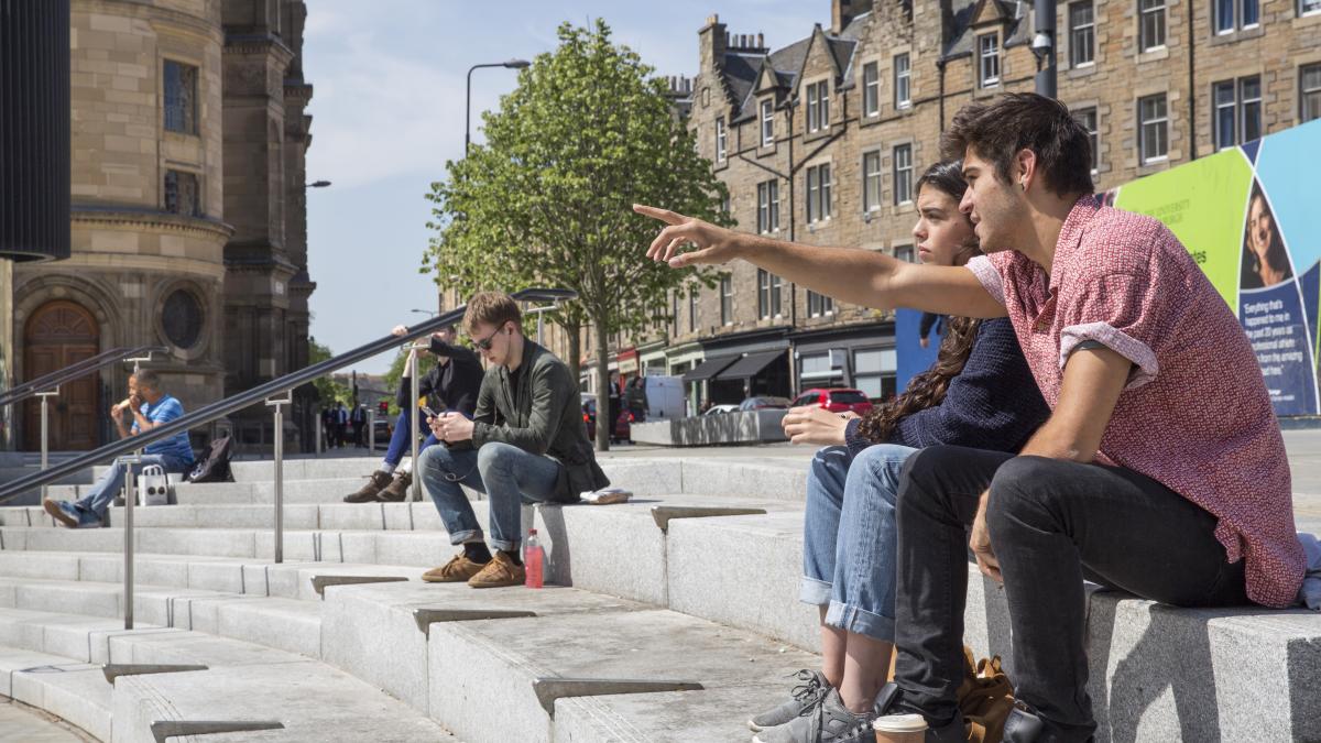 Students sit on the steps on Bristo Square. Some are talking to each other and others are relaxing alone. The image is displayed in landscape orientation in the 16:9 ratio style.