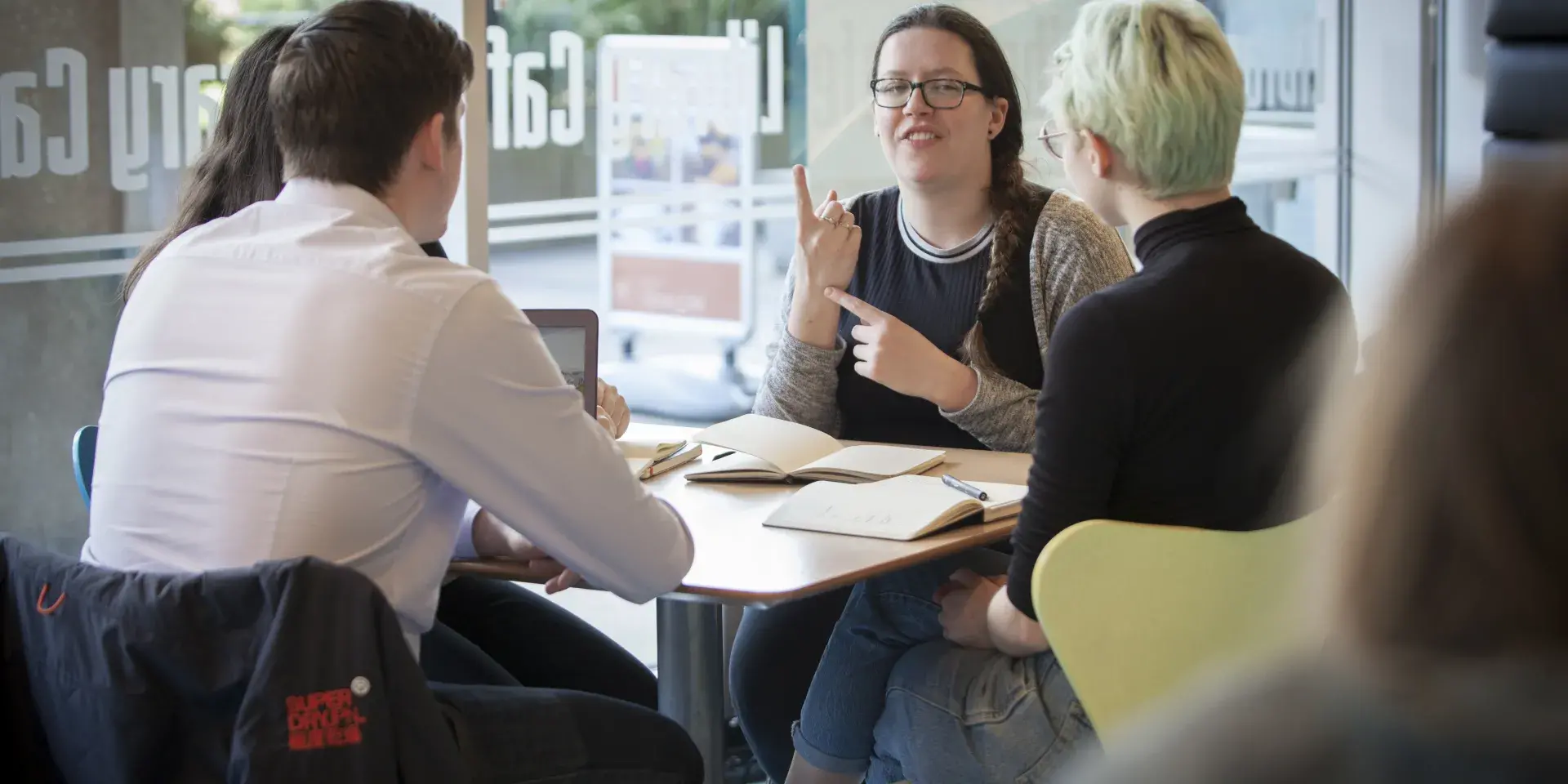 A group of people are sitting around a table in the library cafe, talking to each other using British Sign Language 