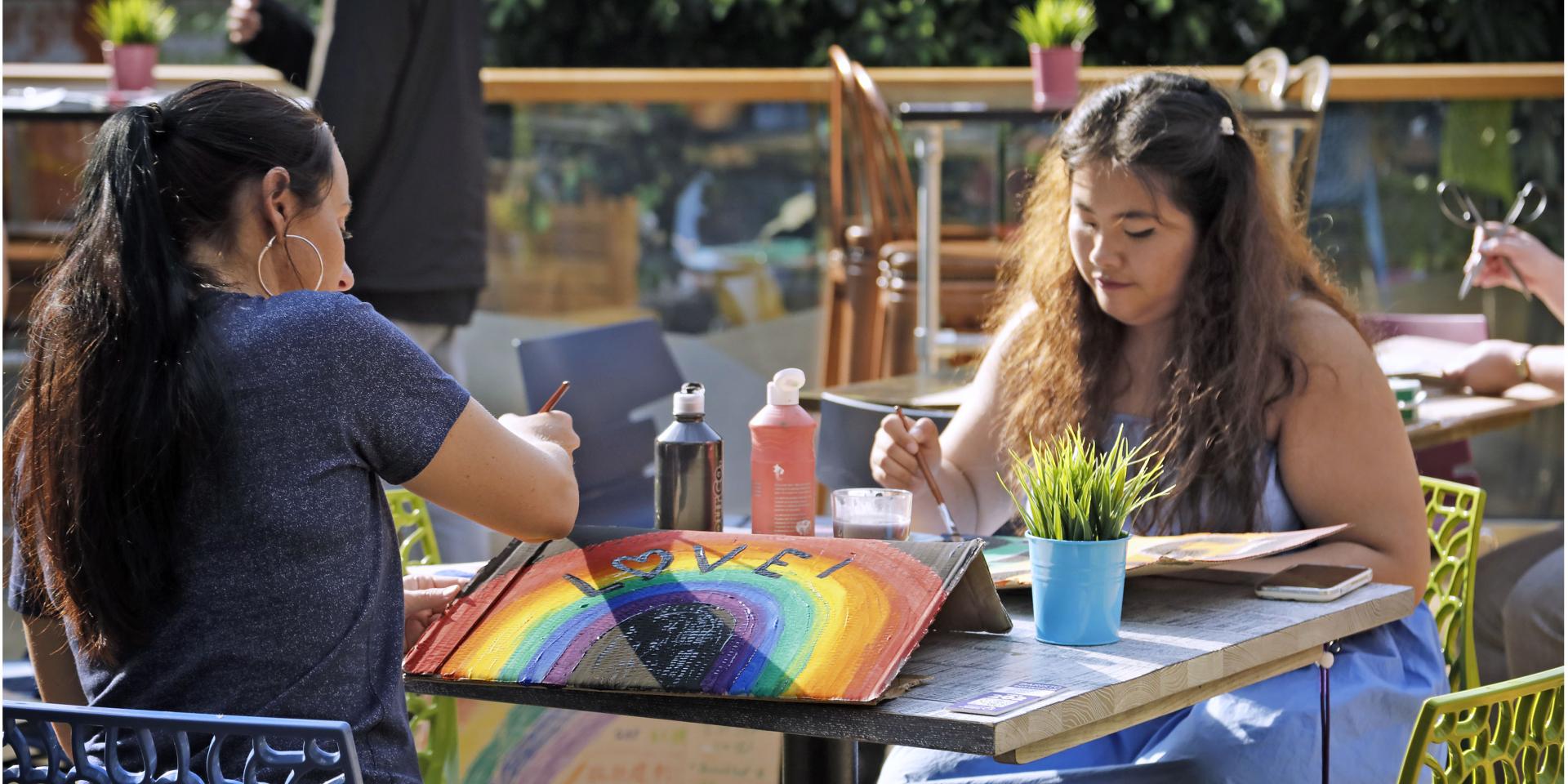 Two women sit outdoors at a table painting rainbow Pride placards, at the University of Edinburgh Pride Event 2023. 