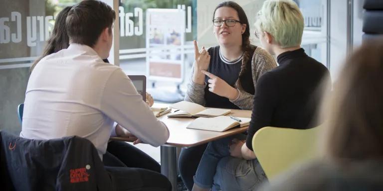 A group of people are sitting around a table in the library cafe, talking to each other using British Sign Language 
