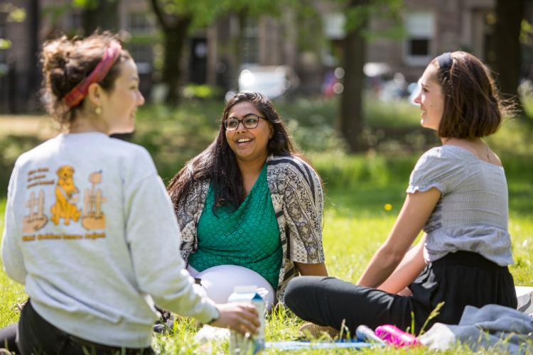 Three young women sit on the grass in the sun in George Square Gardens. They are talking and laughing together. The image is displayed in landscape orientation, with 750x500 pixel dimensions.