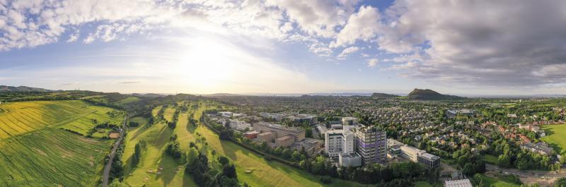 A panoramic view from the air across Edinburgh, with the King's Buildings in the foreground, looking towards Arthur's Seat and Salisbury Crags in the background. The image is displayed in landscape orientation, taking up the full width of the webpage no cropping to the original image..