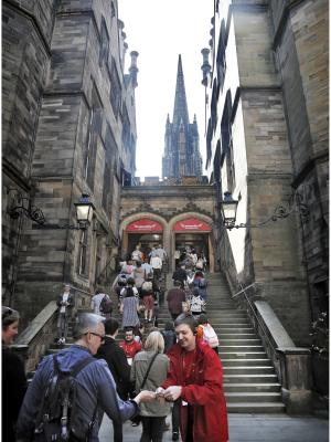 A view up the stairs in front of the historic New College building, with Edinburgh Fringe staff assisting visitors as they enter an events space. The image is displayed in portrait orientation with 300x400 pixel dimensions.