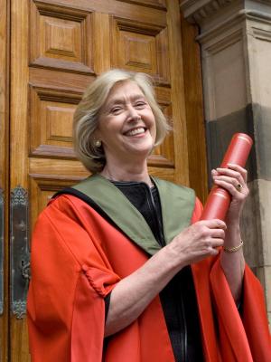 Professor Anneila Sargent stands in red graduation robes in front of one of the doors of McEwan Hall. She is laughing and holding an award scroll.