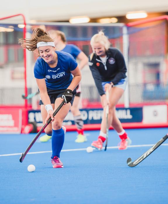 A group of three women play hockey in University brand sports clothes.