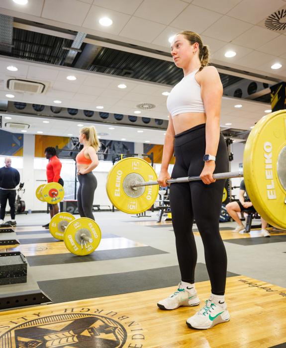 A young woman deadlifts a bar with yellow weights on the end in the Pleasance gym.