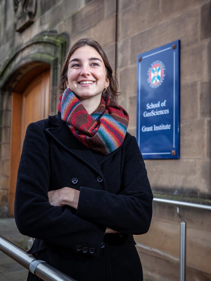 A portrait of University alumna Gail Sant. She is standing in front of the School of Geosciences, smiling at the camera and with crossed arms. The image is displayed in portrait orientation with 675x900 pixel dimensions.